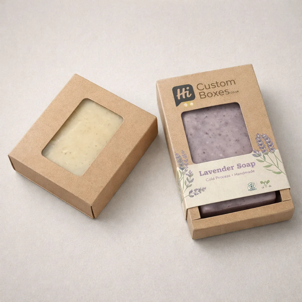 Two brown kraft soap boxes with rectangular cut-out windows, one showing a cream soap bar and one lavender. class=